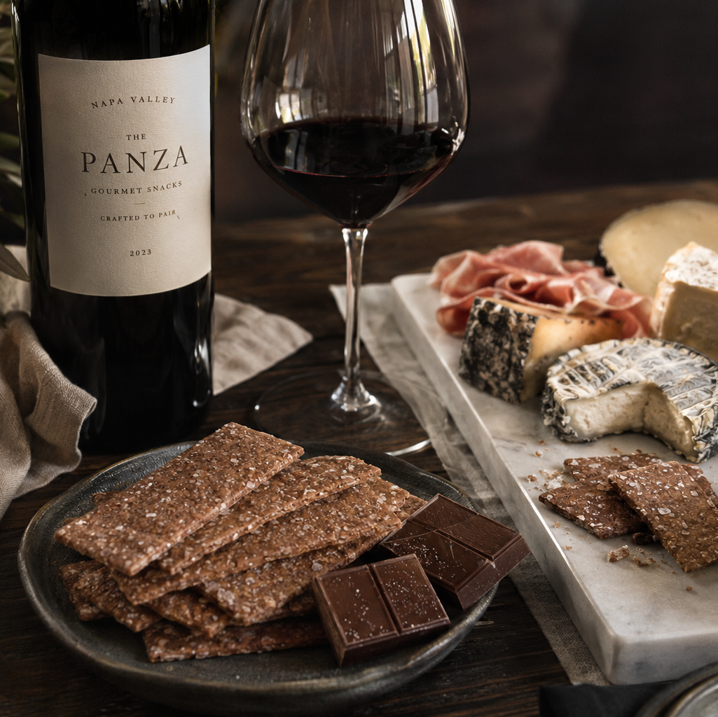 Wine bottle, glass, and charcuterie board with Panza wine on a wooden table and The Panza premium cookies on plate.